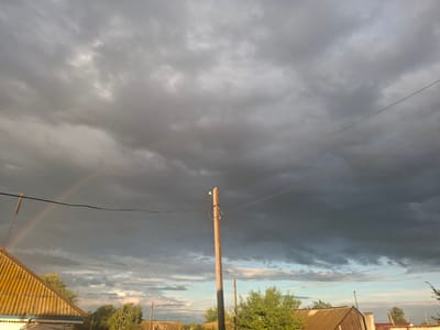 The image depicts a rural landscape with a stormy sky. Dark, heavy clouds dominate the upper portion of the frame, with patches of lighter sky and a hint of a rainbow visible on the left side. In the foreground, a wooden utility pole stands prominently, supporting electrical wires that stretch across the scene. To the left, the yellow corrugated roof of a building is partially visible, and on the right, the rooftops of other structures and green trees can be seen. The overall atmosphere suggests an impending or recent rain shower. The location is identified as Atbasar, Kazakhstan.