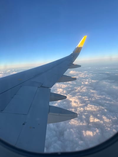 An airplane wing, illuminated by the sun, extends across a vast expanse of clouds, viewed from inside the aircraft. The wingtip, painted bright yellow with "Vueling" logo visible, is tipped upwards against a clear blue sky. The clouds below are fluffy and white, with shadows suggesting sunlight at a low angle, implying either sunrise or sunset. The scene is framed by the curved window of the airplane. The location of Močići, Croatia is not discernible from the image.
