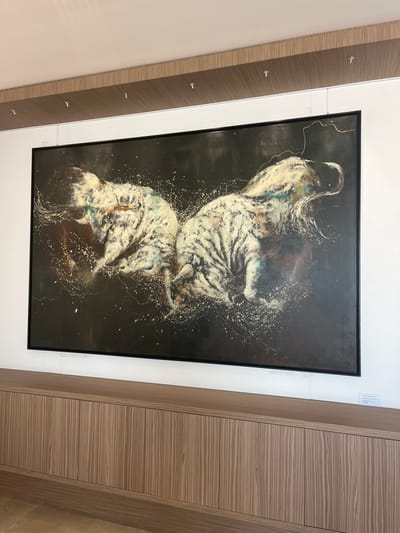 A large, horizontally oriented contemporary painting is displayed on a white wall in an interior space within the city of Galatina, Italy. The artwork, framed in black, features two dynamic, stylized figures resembling elephants, rendered primarily in white, cream, and pale yellow with visible brushstrokes and underlying tones of blue, green, and brown, against a dark, almost black background. White paint splatters emanate from the figures, suggesting movement or energy. A thin, winding gold line is present in the upper right quadrant of the painting. The artwork is suspended by thin wires from silver-colored hooks attached to a horizontal white strip situated below a light-brown wooden panel on the upper wall. Directly beneath the painting, a long, low cabinet or credenza constructed of light-brown wood with distinct vertical grain patterns extends horizontally across the wall. The top surface of the cabinet is flat. A light-colored floor is visible at the base of the image.