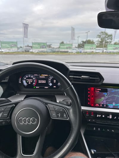 This is a first-person view from inside a car, looking forward through the windshield at a car dealership. The scene is primarily composed of the car's interior: a steering wheel with the Audi logo prominently displayed, a digital dashboard showing speed and navigation information, and a central infotainment screen displaying a map. Various buttons and controls are visible on the steering wheel and dashboard. The digital dashboard shows a speed of 0 km/h, a fuel consumption of 7.7 L/100km, and a time of 20:11. The infotainment screen indicates a navigation route with a destination labeled "Vídeňská" and an estimated arrival time of 20:23. The weather appears overcast and potentially rainy, with water droplets visible on the windshield. Outside the car, rows of vehicles are parked, characteristic of a car dealership lot, with green promotional banners visible in the background. The overall setting is a car dealership on a cloudy day, likely in the early evening, given the time displayed on the dashboard. No people are directly visible in the image.