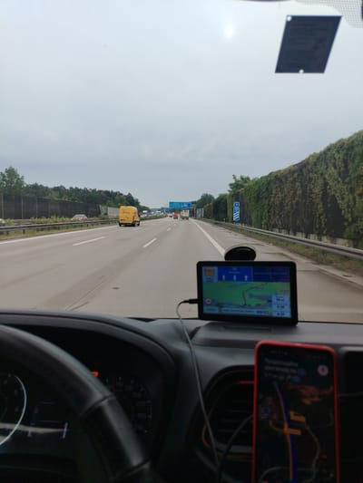 The image is taken from the interior of a vehicle, looking out through the windshield onto a multi-lane highway. The sky is overcast. The highway has at least three lanes visible in the direction of travel. Traffic is present, including a yellow DHL van in the right lane, other cars, and trucks in the distance. On the right side of the highway, there is a dense hedge or ivy-covered barrier that runs along the road. A GPS navigation device is mounted on the dashboard, displaying a map with a route highlighted. A smartphone is also visible in the foreground, also displaying a map application. The dashboard of the vehicle, including a steering wheel and speedometer, is partially visible in the lower part of the image. A sign indicating "9 300 m" is visible on the right side of the highway. The location is identified as being in the city of Königs Wusterhausen, Germany, which is consistent with the presence of a German highway and vehicle markings.