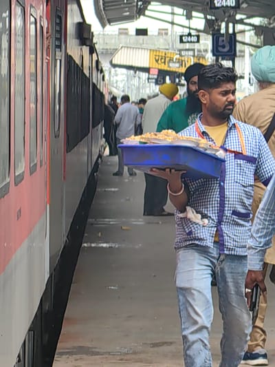 An Indian man in a blue and white checkered shirt and blue jeans walks along a train platform in Khandwa, India, holding a blue plastic tray filled with food. The train, red and white, is on the left side of the image. In the background, people are visible, and signs indicate the station name "Khandwa" and platform numbers. The platform is paved, and there are overhead structures and lighting. The man is looking to his right, and his left hand is on the tray, while his right hand is at his side. The overall scene depicts a busy railway station environment.