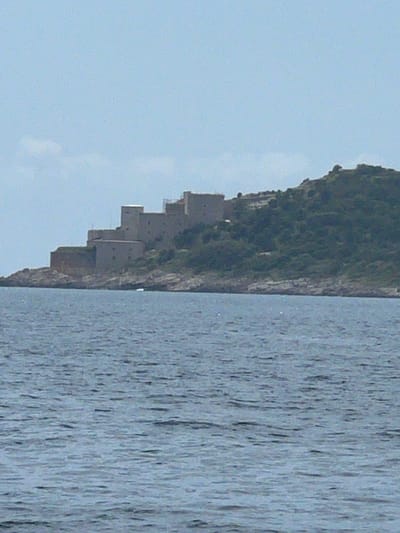 The image presents a view of a terraced structure built into the side of a steep, vegetated cliff in Meljine, Montenegro. The cliff face borders a body of water, likely the Adriatic Sea. The structure consists of several rectangular, block-like levels, constructed from a neutral-toned material, potentially concrete or stone. The water's surface shows ripple patterns. The sky is visible and pale in color.