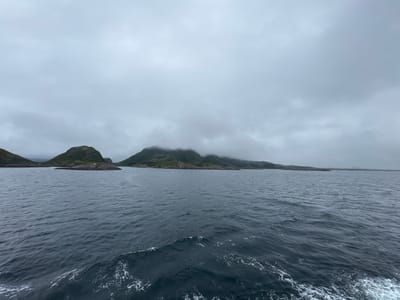 An overcast, wide shot captures the serene waters surrounding Onøya, Norway. A calm, dark blue sea fills the foreground, with gentle waves and white foam indicating movement. In the midground, rugged, green islands with rocky outcrops rise from the water. These islands are partially obscured by a thick, grey mist that clings to their peaks. The background features a faint horizon where the sea meets the sky, which is entirely covered by a dense layer of grey clouds. No people or specific man-made structures are clearly visible, besides a tiny, white building on one of the islands.