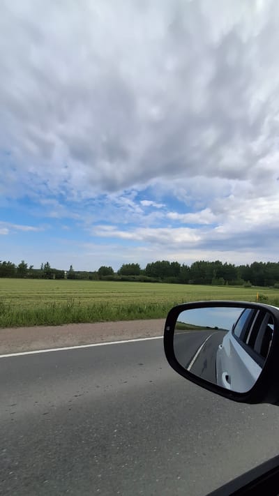 The image presents a daytime landscape view from a moving vehicle, looking out towards the side. A dark asphalt road, with a solid white line marking its edge, occupies the right side and foreground of the scene. To the left of the road, a wide expanse of vibrant green grass or field stretches horizontally, bordered by a dense line of tall, dark green trees forming the distant horizon.
Above the tree line, the sky is partly cloudy, displaying a mix of bright white and soft grey clouds, with noticeable patches of clear blue sky peeking through. In the upper right corner, a black car side mirror is prominent, reflecting a segment of the road ahead, the blue sky, and a white section of the car's own body.
The scene depicts travel or a journey along a rural or intercity road, consistent with being in Annino, Russia. The weather appears to be mild and partly overcast. No discernible text is visible within the image.