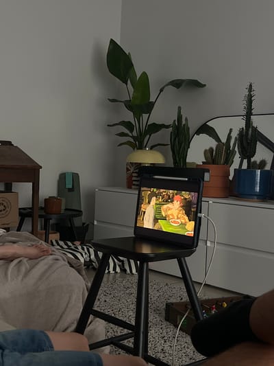 The image shows an indoor scene, likely a bedroom or living room, with a tablet displaying an animated scene placed on a black stool. The tablet is connected to a white cable. In the foreground, a person's legs and feet are visible, with one foot resting on a beige blanket. A portion of another person's legs, clad in denim, is also visible. Behind the stool, a small foosball table is placed on a grey, textured rug. The room is decorated with several potted plants, including a tall Bird of Paradise plant and various types of cacti. A dresser with multiple drawers is visible against the wall, along with a wooden table and a black side table with small potted plants. The lighting suggests it is evening or the room is dimly lit. The image is devoid of any specific clues that would indicate the location within Neuville-en-Condroz, Belgium.