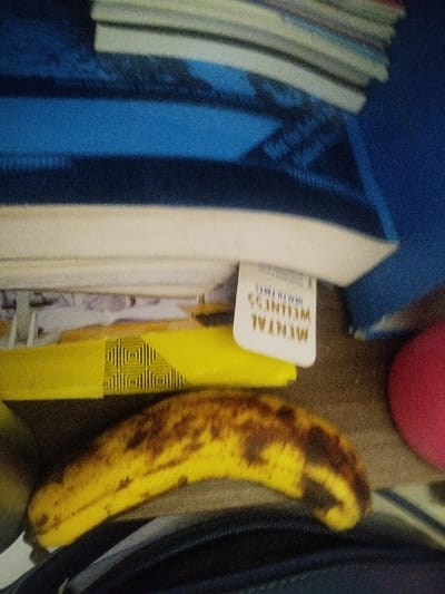 The image depicts an indoor scene in Keekonyokie ward, Kenya, presenting a collection of stationary objects arranged on a dark, flat surface. In the foreground, a portion of a dark blue or black zippered bag or case is visible at the bottom. Centrally, one ripe banana, predominantly yellow with numerous brown spots and patches indicating advanced ripeness, rests horizontally. To the left of the banana, a yellow object, possibly a book cover or folder, features a black geometric pattern. Above this yellow object, a white tag or bookmark is partially visible, inverted, displaying the text "MENTAL WELLNESS HEALTH FACTS." In the background, a stack of books or papers with white spine edges is present, resting on a blue surface, likely a book or fabric. To the far right, a segment of a red spherical or rounded object is visible. The scene is static, with no human or animal presence.