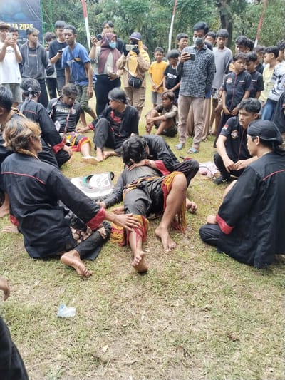 The image depicts an outdoor traditional or cultural event in Puro, Indonesia, featuring participants and a crowd of onlookers. In the foreground, several individuals are seated on a grassy patch. Two central figures, dressed in black garments with red accents and leg wraps with yellow and red fringes, appear disoriented or exhausted. One lies on their side, covered in mud or dirt, while another person attends to them, gently touching their arm. A decorative prop resembling a horse's head or mask lies on the ground between them.

Surrounding these central figures, a crowd of people observes the scene. Many standing onlookers, mostly young men, are actively recording the event with their smartphones. They are dressed in casual attire, such as t-shirts, hoodies, and baseball caps. A woman wearing a hijab and a face mask is also among those recording. The background consists of lush green trees and a partially visible banner with text such as "TA 2025" and "agen, 22 Ar".

The ground is grassy with some muddy patches. A discarded plastic bottle is visible on the grass. The event appears to be taking place during daylight hours, possibly under an overcast sky, given the soft lighting. The expressions of the central figures suggest a state of trance or extreme fatigue, while the attendees show concern, and the audience exhibits curiosity and engagement. Other visible text on clothing includes "SPEDIES BRUTR PREDAT" and "BROKEN ANGEL".