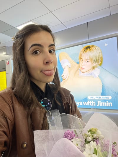 The image shows a woman in Goyang-si, South Korea, standing in what appears to be a public space, likely an airport or train station, based on the background. She is holding a bouquet of flowers wrapped in white netting. Behind her is a large advertisement featuring an image of the South Korean celebrity Jimin, with the text "ALL FOR JIMIN" and "Days with Jimin" visible. The woman is wearing a brown leather jacket and a black top underneath. A pair of round sunglasses are tucked into the collar of her jacket. She has brown hair and is looking at the camera with a slight smile and her tongue sticking out slightly. A small gold-colored flower hair clip is visible in her hair. The background is a modern, tiled interior with a yellow sign near the bottom of the frame.