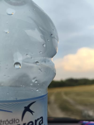 A close-up, side-view, outdoor, waist-up shot shows a clear plastic water bottle with condensation droplets on its surface. The bottle is partially filled with water. In the background, a blurred landscape features a field, trees, and a cloudy sky. The bottom portion of the bottle displays a label with blue and white patterns and the silhouette of a bird. The scene is set in Stanisławowo, Poland.