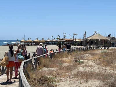 This is a wide-angle, eye-level, daytime, outdoor photograph taken on a sunny day in San Roque, Spain. The scene depicts a wooden boardwalk path winding through a sandy beach area. Several people are walking on the boardwalk, appearing to be heading towards or away from the beach. The beach itself is visible in the background, with numerous thatched-roof umbrellas and some buildings, including a prominent wooden gazebo-like structure. The ocean and a clear blue sky are visible beyond the beach. The vegetation in the foreground and along the sides of the boardwalk is dry and sparse, typical of a coastal environment in warm weather.
