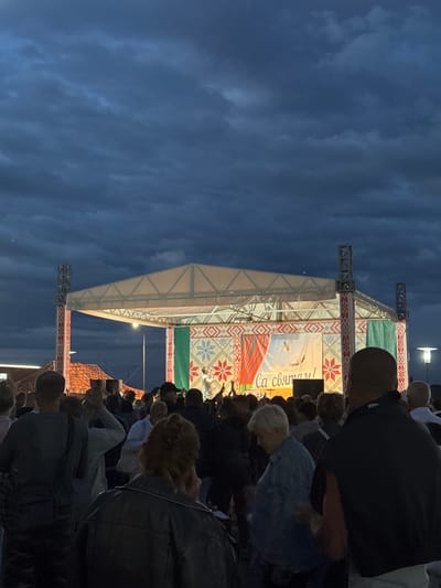The image depicts an outdoor evening event in Uzda, Belarus, under a cloudy, dark sky. A crowd of people is gathered in front of a stage adorned with traditional Belarusian patterns and flags. On the stage, a performer is visible, possibly singing or speaking, illuminated by stage lights. The backdrop of the stage features a banner with Cyrillic text that appears to read "Са святам!" which translates to "Happy Holiday!". The overall atmosphere suggests a festive gathering, likely a local celebration or concert.