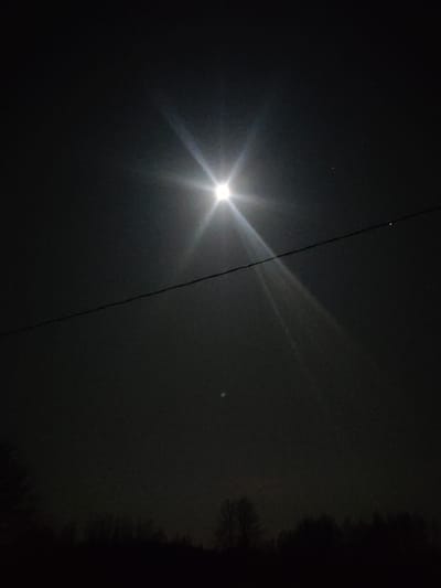 The image captures a nighttime outdoor scene in Jaunaglona, Latvia. A singular, intensely bright celestial object, consistent with the full or near-full moon, is positioned in the upper central sky, emitting a prominent starburst lens flare. A thin, dark utility line traverses horizontally across the mid-ground of the frame. Below this line, to the left of center, a small, faintly luminous, circular object is discernible against the dark sky. The lower quarter of the image is occupied by the silhouetted profile of a dense treeline or distant vegetation. The overall environment is dark, indicative of night, with minimal ambient light. No individuals, specific actions, or interactions are visible.