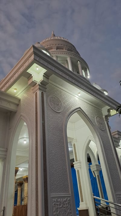 This is a low-angle, full-shot, outdoor photograph of the exterior of a mosque in Lhokseumawe Teungoh, Indonesia, captured at dusk. The mosque is predominantly white with ornate architectural details. A prominent dome is visible at the top of the structure, surrounded by columns and decorative moldings. The facade features arched doorways and intricate relief carvings on its walls. Several lights are illuminated on the exterior, casting a warm glow against the twilight sky, which is filled with diffused clouds. Inside the arches, the blue tarping suggests ongoing construction or renovation. The scene appears serene and majestic, highlighting the mosque's grandeur against the fading light. There are no people visible in the frame.