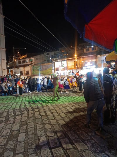 The image captures a night scene in the city of El Cisne, Ecuador, featuring a cultural celebration in a town square paved with cobblestones. A crowd of people, varying in age and attire, are watching a performance. Some are seated on the left side of the frame. Several individuals are dressed in traditional clothing. A costumed performer is actively moving through the illuminated square. Overhead, power lines stretch across the scene. A large umbrella-like structure with red and blue fabric is positioned at the upper right of the image. Buildings with lights visible in windows form the backdrop of the event. A person in the right corner is using a cell phone to record the festivities.