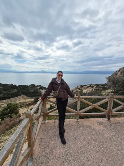A female subject stands centrally on a reddish-brown gravel observation deck located in the Municipal Unit of Loutraki - Perachora, Greece. She faces the viewer, wearing a dark brown puffer jacket, a light grey turtleneck, black trousers, and black boots. A light brown crossbody bag is slung over her left shoulder, and she wears dark sunglasses. Her arms rest on a light brown wooden railing with a criss-cross pattern, which spans the foreground. Beyond the railing, a steep, rocky, vegetated slope with sparse green and dry foliage descends towards a large body of water, indicative of a bay or gulf. Distant landmasses, appearing as hills or mountains, are visible across the water on the horizon. The sky is predominantly overcast with grey clouds.