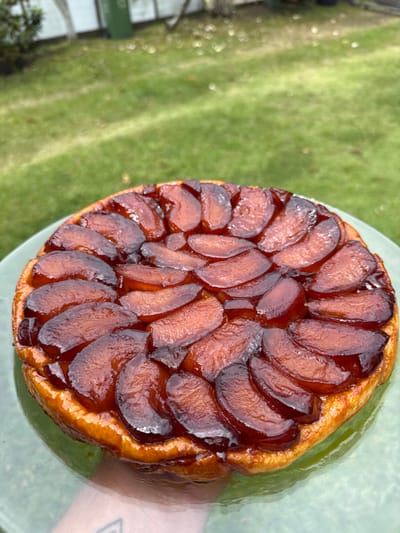 A circular Tarte Tatin is centrally positioned on a light green, possibly glass, plate. The tart features a golden-brown, flaky pastry crust supporting a dense arrangement of caramelized apple slices. These slices are dark amber to reddish-brown, glossy from the caramelization, and layered in a concentric pattern. A human hand, with a faint tattoo visible on the inner wrist, supports the plate from below. The background is an out-of-focus outdoor environment, dominated by bright green grass. In the upper left portion of the background, a dark green cylindrical bin and a light-colored fence or wall are partially discernible. The scene is located in Māhina, France.