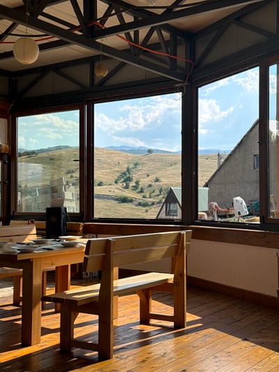 An interior view of a rustic dining area with large windows overlooks a sun-drenched, rolling landscape. The room features wooden tables and benches, with sunlight casting sharp shadows across the polished wooden floorboards. In the foreground, a wooden table is set with plates and cutlery. Beyond the table, a wooden bench with slatted seating and backrest is visible. The expansive windows offer a view of a dry, grassy hillside dotted with trees and a few buildings. In the middle distance, a small, dark-colored speaker sits on the table. The ceiling is a mix of exposed metal beams and wooden panels, with two paper lanterns hanging from above. The scene appears to be located in Rudinice, Montenegro.