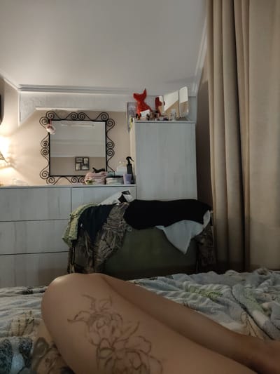 The image depicts a bedroom scene. In the foreground, the lower body of a person is visible, lying on a bed covered with a textured blanket. A tattoo of a floral design is visible on the person's leg. In the background, there is a dresser with several drawers, a mirror with an ornate frame hanging above it, and a tall white cabinet. On top of the cabinet, there are some decorative items and toiletries, including a red cat-shaped figurine and several bottles. A dark curtain hangs on the right side of the frame. The scene appears to be indoors, possibly in a residential setting. The lighting suggests it might be evening or nighttime. There is no visible text in the image.