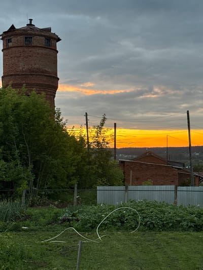 This image captures an exterior scene at dusk, likely in or near the city of Malmyzh, Russia. Dominating the left side of the frame is a tall, weathered brick water tower, its cylindrical structure showing signs of disrepair with broken windows and a damaged roof. A dense canopy of green trees partially obscures the tower, reaching from the foreground towards its base.
In the background, a vibrant sunset paints the sky with intense shades of orange and yellow, with softer hues of pink and gray clouds interspersed. Silhouetted against this dramatic sky are power lines supported by wooden poles, suggesting the presence of infrastructure. Below the sunset, a line of dark trees forms the horizon, with glimpses of buildings in the distance.
Closer to the viewer, a red brick building with a dark roof is visible. In the foreground, a grassy area transitions into a garden or cultivated plot with rows of green plants, possibly potatoes, exhibiting small white flowers. A light-colored hose is coiled on the ground in the garden. A simple fence, constructed from wooden posts and corrugated metal sheeting, runs across the mid-ground, separating the garden from the area with the brick building. The overall atmosphere is tranquil and rural, with the natural beauty of the sunset contrasting with the rustic, somewhat neglected man-made structures. There are no people visible in the scene.