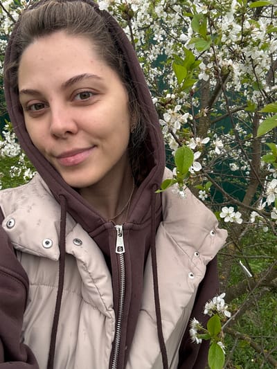 A young woman is posing for a photo. She is wearing a brown hooded sweatshirt and a beige quilted jacket. The jacket is partially zipped up. She is standing outside, with a tree full of white blossoms in the background. The leaves of the tree are green. The background appears to be a fenced yard. There is no indication that the location is in the city of Nikulino, Russia.