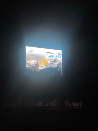 The scene depicts a brightly illuminated rectangular billboard at night in Dabouq, Jordan. The majority of the image is dark, with the billboard serving as the primary light source. In the lower portion of the image, dark silhouettes of trees or dense foliage are visible, partially obscuring the bottom edge of the billboard.
The billboard itself features an advertisement for "JERUSALEM INSURANCE" (القدس للتأمين), with their logo—a stylized archway or building—positioned in the upper left. The main Arabic text prominently asks, "عملت حادث و ع كروكافوبيا؟" (Had an accident and Crocophobia?), followed by "حلّه مع تأمين القدس الشامل" (Solve it with comprehensive Al-Quds Insurance). An illustrative graphic occupies the center-left, showing a distressed, red, oval-shaped cartoon character with white eyes and a blue mouth, appearing submerged or entangled in a pile of various objects or debris. A phone number, "06 4040..." (partially visible), is displayed in the bottom right corner of the billboard. No individuals are present in the visible frame, and no specific actions or interactions are depicted.