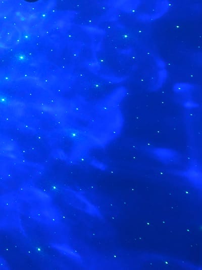 The image is a vertical, close-up shot of a starry night sky projected onto a surface, likely a ceiling or wall. The dominant color is a deep, vibrant blue, simulating the night sky. Scattered across this blue expanse are numerous small, bright white and greenish-white dots, representing stars. Some of these stars appear more luminous and slightly larger than others, suggesting variations in brightness or proximity. There are also softer, cloud-like smudges of lighter blue interspersed with the stars, creating a nebula or galactic effect. The lighting is artificial, with the projection creating a magical, ethereal atmosphere. There are no people, animals, or identifiable objects in the scene other than the projected starry pattern. The image does not provide any visual cues that would allow for the identification of its location within Calfa, Moldova.