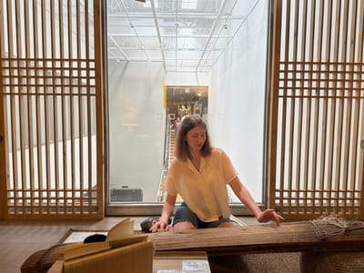 A young woman with shoulder-length brown hair is sitting on the floor in front of a traditional wooden instrument, possibly a gayageum, with her right hand outstretched, touching its strings. She is wearing a short-sleeved white blouse and denim shorts. The instrument is placed on a woven mat. To her left is a wooden bookstand with an open book. The scene is set in a modern building with large windows and a white industrial ceiling structure visible through them. Outside the windows, a staircase with people can be seen. The surrounding area is framed by intricately carved wooden panels, resembling a traditional Korean screen or door. The overall environment suggests an indoor cultural space or exhibition within Goyang-si, South Korea.