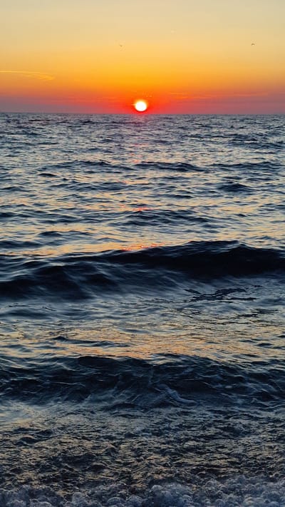 The image depicts a sunset over the Adriatic Sea in Zadar, Croatia. The sun, a vibrant orb of red and orange, is low on the horizon, casting a warm glow across the water and sky. The sea is rough, with waves creating a textured, almost abstract pattern of light and shadow. The water's surface is a mix of deep blues and grays, reflecting the dramatic colors of the sunset. Along the left side of the frame, the sky transitions from a pale yellow to a fiery orange as it approaches the sun. The overall mood is serene and awe-inspiring, capturing the beauty of a Mediterranean sunset. There are no people or objects visible in the scene, emphasizing the natural grandeur of the landscape. The image appears to be a photograph, possibly taken from a boat or a coastal vantage point, offering a dynamic perspective of the churning waves. The time of day is clearly sunset, and the weather seems clear, with the sun prominently displayed.