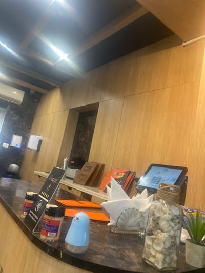 An interior view depicts a counter area within a commercial establishment in Monastir, Tunisia. The counter is dark, likely marble, and extends across the lower portion of the frame. On the counter, from left to right, are two clear plastic containers of "TOOTHPICK" brand toothpicks, followed by a transparent acrylic display stand prompting Google reviews with a 5-star rating and an NFC symbol for contactless feedback, including French text. Adjacent is a light blue, bird-shaped automatic hand sanitizer dispenser. Further right, stacks of menus, some brown and some orange/red, are visible behind a clear glass napkin holder containing white folded napkins. A clear glass jar filled with small, light-colored seashells, tied with twine, stands next to a small potted green plant in a grey pot. To the far right, a black point-of-sale (POS) system with a tilted touchscreen displaying a grid interface is positioned.
The wall behind the counter is paneled with light-colored wood, featuring horizontal and vertical seams. To the left, this wood paneling transitions to a darker, possibly marble-patterned wall section. Mounted on the dark wall is a white air conditioning unit, and below it, two white wall-mounted dispensers (one with a blue light, likely for hand sanitizer, and another for paper towels) are visible above a dark, cylindrical trash receptacle. A recessed rectangular niche within the wood-paneled wall also features a dark, marble-like interior. The ceiling is dark with recessed lighting fixtures and lighter wood beams forming a grid pattern, producing visible lens flares. No individuals are present.