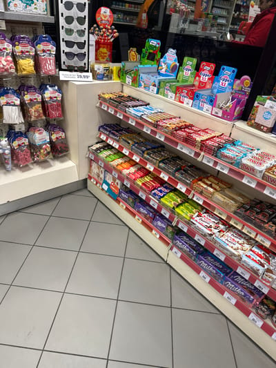 A well-stocked convenience store aisle is pictured with shelves overflowing with candy and snacks. Dipper XL and Chupa Chups lollipops sit alongside Milka and Kinder Bueno chocolate bars. Lower shelves display bagged candies, while sunglasses are displayed at eye-level with a price tag of 19.99. The floor is tiled in a light gray grid pattern.