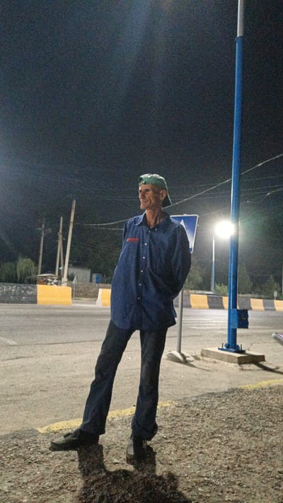 The image shows a man standing outdoors at night, near a road. He is wearing a dark blue button-down shirt, dark pants, and a green baseball cap. His hands are behind his back. The background includes a street, yellow and black barriers, streetlights, power lines, and a blue pole with a sign. The man is casting a shadow on the ground. The location is identified as Pistamazor, Tajikistan.