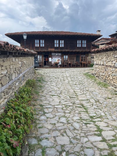 The image shows a cobblestone street leading towards a traditional wooden building in the city of Zheravna, Bulgaria. The building, likely a mehana (tavern), has a dark wood facade with multiple windows and a red tile roof. A sign is visible on the front of the building, featuring Cyrillic script. Stone walls flank the street on either side. There are several tables and chairs set up outside the building, suggesting an outdoor dining area. A few people are visible near the entrance of the building and seated at one of the tables. The sky is overcast, and there is some greenery growing along the stone walls and between the cobblestones.