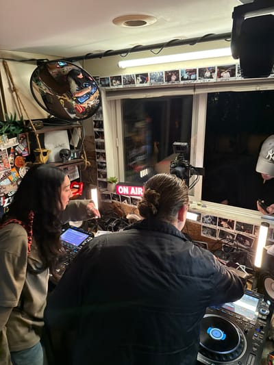 Inside a room, two people are present; one stands wearing a black jacket and the other sits. A DJ mixer is visible near the standing individual. A window is adorned with photos, and an "ON AIR" sign is lit. A camera is mounted near the window. Various stickers cover the lower portion of the room. A convex mirror is mounted on the wall.