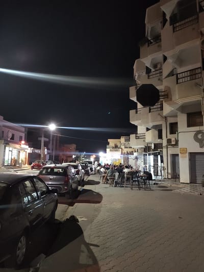 The image captures a nighttime street scene in an urban environment. A multi-story building, likely an apartment complex or hotel, dominates the right side of the frame. The building is white with balconies and dark railings, and features some windows and air conditioning units. In front of the building, an outdoor seating area with tables and chairs is occupied by several people, suggesting a cafe or restaurant.
To the left of the building, the street is lined with cars parked along the side. Beyond the parked cars, there are storefronts, one of which is labeled "LE PANIER FIN". Streetlights illuminate the scene, creating long streaks of light across the image. The ground is paved with interlocking bricks. The sky is dark, indicating it is nighttime. The overall impression is a lively street scene in a Tunisian city, with people enjoying an evening out.