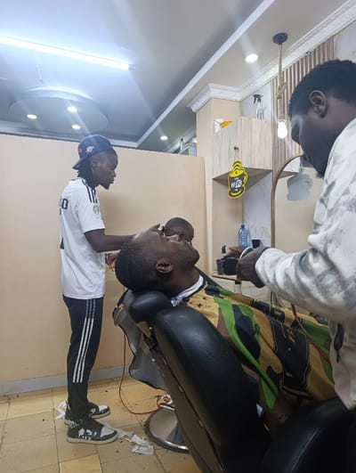 Inside a barbershop, two barbers are working on a customer seated in a black barber's chair. The customer is draped with a camo-patterned cape. One barber stands to the left wearing a white soccer jersey, black pants, and a cap. He is focused on the customer's hair with clippers. Another barber stands to the right, also holding clippers. The shop has bright overhead lighting and a modern decor, including a "WiFi" sign shaped like a coffee cup. Scraps of hair litter the floor.