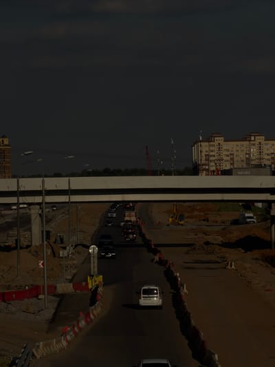 The image depicts a construction site on an overcast day in Novodrozhzhino, Russia. In the foreground, a white van is parked on a road, with the license plate partially visible. The road is lined with temporary traffic barriers, some of which are red and white. Beyond the barriers, construction equipment, including a yellow excavator, is present on a dirt surface. In the middle ground, a highway overpass stretches across the scene, supported by concrete pillars. Traffic is visible on the road beneath the overpass, with several cars and trucks. In the background, a multi-story residential building with cream-colored walls and numerous windows stands, with the word "ФИТНЕС" (Fitness) visible on its facade. The sky is dark and cloudy, suggesting a late afternoon or evening setting.