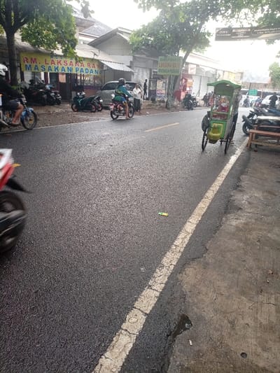 The scene is a street in Kecamatan Caringin, Indonesia. The street is wet, indicating that it may have rained recently. There is a white painted line dividing the asphalt road. There are a few parked motorcycles on the side of the road and a few people are walking around. A food cart is parked on the right side of the road. There is a yellow sign above a store, which reads "RM. LAKSANA RAY MASAKAN PADANG". There is another yellow sign above another store, which reads "RUMAH MAKAN LAKSANA RAY MASAKAN PADANG". There are some buildings on the left side of the road. There is a tree in the foreground and some trees in the background. The buildings and shops are all located along the street. There are a few people standing on the sidewalk.  There is a motorcyclist riding on the road, passing a food cart on the right.  A few people are walking along the sidewalk in the background.  There is a piece of litter on the road.