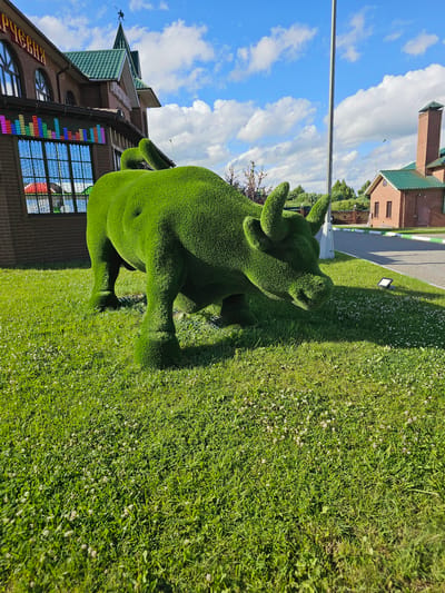 This image depicts an outdoor scene in the city of Ilyinskoye, Russia, on a sunny day with a partly cloudy blue sky. In the foreground, a large, artificial bull sculpture covered in green artificial grass stands on a lush green lawn dotted with small white flowers. The bull is realistically sculpted, with visible legs, body, head, and horns. To the left of the bull, a brick building with arched windows and a green tiled roof is visible. One window has a colorful LED display resembling an equalizer. To the right, a paved pathway borders the lawn, and further back, another brick building with a green roof and a chimney is present, along with some trees and a fence. A tall, slender pole stands near the bull sculpture, and a spotlight is visible on the pathway.