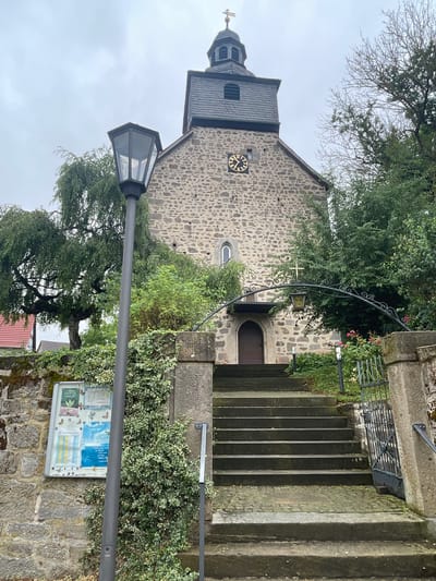 The image shows the exterior of the Evangelical Church in the municipality of Ottrau, Germany. The church is constructed of stone, with a tall tower topped by a dark-pitched roof and a weather vane. A clock face is visible on the tower. In front of the church is a set of stone stairs leading up to the entrance. A wrought-iron archway with a lantern hangs over the entrance. The area is surrounded by greenery, including trees and bushes, and a stone wall with a notice board. A street lamp stands prominently in the foreground. The sky is overcast.