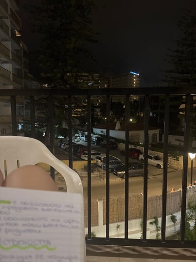 The image is a nighttime view from a balcony overlooking a street lined with parked cars and buildings in Fuengirola, Spain. A white plastic chair is partially visible in the foreground, with a blurred notebook resting on the lap of an unseen person. The notebook contains handwritten text. Beyond the balcony railing, several vehicles are parked on the street, including a dark grey sedan, a white hatchback, and a red car. A white building with a red-tiled roof and a chimney is visible, as is a larger hotel with illuminated windows and a neon sign that reads "HOTEL". Tall pine trees are scattered throughout the scene, and a lamppost is visible on the right side of the frame. Two individuals can be seen standing near a fence in the background on the right. The overall impression is a residential or tourist area at night.