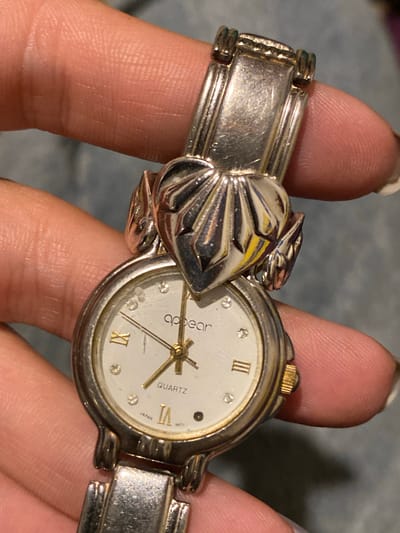 A person is holding a silver-colored watch with a white face. The watch features Roman numerals and the brand name "Appear Quartz" on the dial. The band is made of silver-colored metal and includes a decorative, shell-shaped element. The watch appears to be worn. The background is a blurred, neutral tone. The location in Monte Grande, Argentina, cannot be determined from the image.
