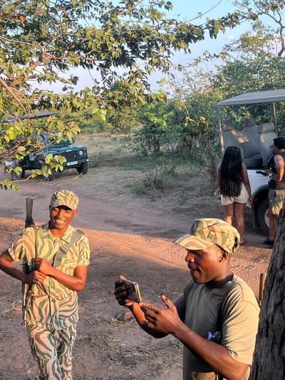 In Victoria Falls, Zimbabwe, the scene features two male African park rangers in camouflage uniforms. One ranger is holding a rifle and smiling, while the other is looking at a cell phone. In the background, there is a group of people next to a safari vehicle and a green SUV. The surroundings include trees and dry dirt.