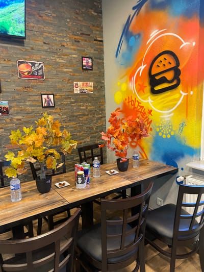 This image depicts the interior of a casual dining establishment located in Holtzheim, France. The scene focuses on a dining area featuring a long, rustic wooden table and several dark brown wooden chairs with black padded seats. The table is set with two artificial fall foliage arrangements in black pots—one displaying yellow and green leaves, the other orange and red leaves. On the table, various beverage items are present, including multiple soda cans (red, silver, blue), two plastic bottles of mineral water, and a black and yellow paper cup. Four small white square plates, each holding a single brown food item, are also distributed on the table.

The wall to the left is clad with irregular grey stone veneer and adorned with several items: a wall-mounted television displaying green and blue imagery, a metal sign depicting a burger with "HOT BURGERS FAST FOOD" text, a metal sign featuring a classic car on "Route 66 ARIZONA USA," and two small framed pictures, one depicting a person and another a figure, possibly a zombie or monster. The adjacent wall, to the right, is painted with vibrant graffiti art. This mural includes a large black outlined burger icon, various spray paint splatters and strokes in orange, yellow, blue, and white, and the word "HOLTZHEIM" rendered in red and black spray paint. A white radiator is visible on the lower section of the graffiti wall, beneath a window sill. No individuals are present in the scene.