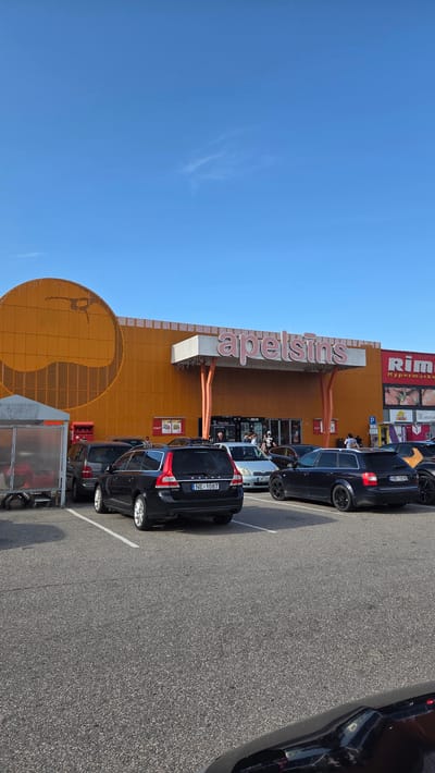 An exterior, eye-level, vertical shot shows the "Apelsins" supermarket in Stapriņi, Latvia, under a clear blue sky with a few wispy clouds. The supermarket building is primarily orange with large, pink lettering that reads "apelsins". To the left, a large, circular orange design is visible on the building's facade. The entrance area is marked by orange poles supporting a white overhang. To the right of the entrance, signage for "Rimi Hypermarket" is present. The foreground is dominated by a black asphalt parking lot with white parking lines. Several cars are parked in the lot, including a black Volvo station wagon in the center with license plate "NE-1087", a silver sedan, a dark blue Audi station wagon with license plate "HB-1010", and other vehicles partially visible. A grey trolley shelter with red elements is situated on the far left of the image. A few pedestrians are visible near the supermarket entrance. The overall scene depicts a typical day at a retail establishment with a parking lot.