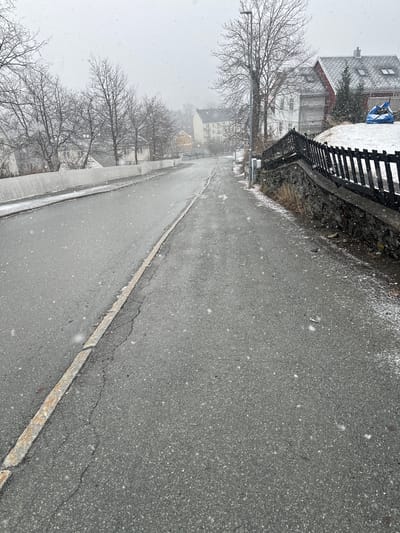 The image shows a street in Trondheim, Norway, during a light snowfall. The road surface is asphalt, showing some cracks and a concrete curb on the left. On the right side of the road is a low wall, seemingly made of stone and topped with a dark-colored wooden fence. Several bare trees are visible along the left side of the street. Buildings are visible in the background. The sky is overcast, indicative of a snowy or wintery weather.