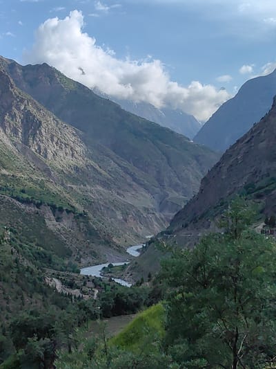 The image depicts a mountainous landscape with a river flowing through a valley. The terrain is characterized by steep, rocky slopes covered in sparse vegetation and patches of green. In the foreground, dense trees partially obscure the view. The river, appearing wide and light-colored, meanders through the valley floor, surrounded by a winding road and some scattered buildings. The sky above is blue with scattered white clouds, and distant snow-capped mountains are visible. The scene suggests a remote, natural environment, likely in a mountainous region of India, given the mention of Kardang.
