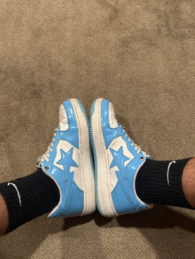 A pair of light blue and white patent leather sneakers with a star design is shown from a top-down perspective, resting on a brown carpet. The sneakers are positioned in a V-shape, with the toes pointing outward. Both shoes feature white laces and white soles, with a blue rubber outsole. The left shoe displays a light blue star outlined in white, while the right shoe shows the same design on its outer side. Black socks with a white Nike swoosh logo are visible on both feet, peeking out from beneath the shoes. The overall environment appears to be indoors, on a carpeted floor. The location is identified as Ocean City, United States of America, though no specific landmarks or context within the city are provided by the image itself.