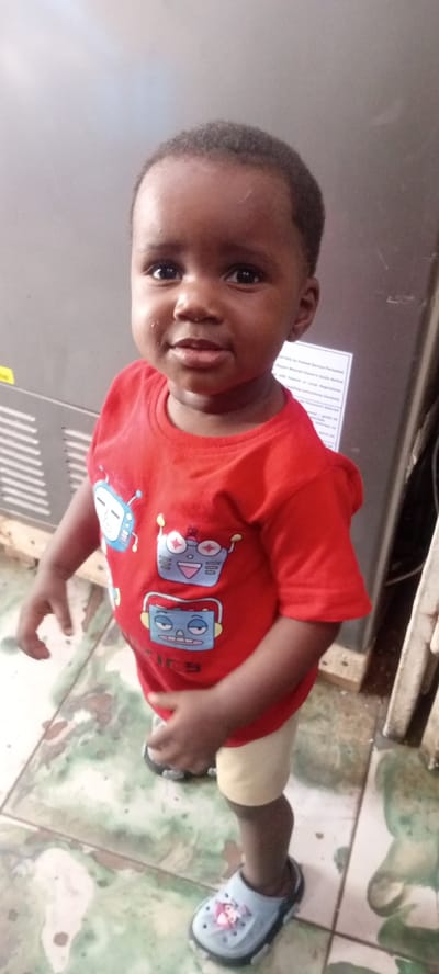 A waist-up portrait features a young child, likely a toddler, standing indoors and looking directly at the camera with a gentle, slight smile. The child has dark skin, a round face, and short, dark, closely-cropped hair. Beads of sweat or moisture are visible on the child's forehead, cheeks, and upper lip.
The child is dressed in a vibrant red short-sleeved t-shirt adorned with light blue cartoon robot designs. Text that appears to be "ICY" or "TICY" is partially visible below one of the robots. They wear light-colored shorts, possibly beige, and light blue slip-on shoes resembling Crocs. One shoe displays a printed character that looks like a Disney princess, possibly Elsa.
The setting appears to be a utilitarian indoor space. Behind the child is a large, dark gray or black rectangular object, possibly an industrial refrigerator or appliance. A light yellow sticker is visible on its left side. On the right, a white sheet of paper with black text is attached, displaying phrases such as: "NOT ONLY to Trained Service Personnel," "Report Unusual/Illegal Behavior," "This unit complies with Federal or Local Regulations," "Reading instructions carefully," "Maximum Performance Achieved," and "do not put anything inside" (partially visible).
The floor is covered with light-colored tiles, possibly white or cream, interspersed with irregular green and brown patterns. The image is brightly lit, suggesting daytime. The location context provided is Kwachiri, Nigeria.