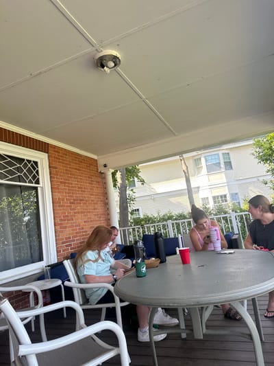 Four individuals are seated at a grey, oval outdoor table on a wooden deck, likely a porch or patio. The scene appears to be in Harbor Springs, USA, with a residential building visible in the background. One woman with long red hair, wearing a light blue striped short-sleeved shirt, is sitting at the table, looking down at her phone. Beside her, a man wearing a white shirt is also seated at the table, looking down. To the right, two other women are seated. One is wearing a pink tank top and looking at her phone, while the other, wearing a black t-shirt, is facing her. On the table, there are several items, including water bottles, a red cup, and what appears to be a small container of food. The porch ceiling has a visible light fixture, and brickwork is present on the left side of the image, suggesting the architecture of the building. The overall atmosphere is casual, possibly a gathering or meal.